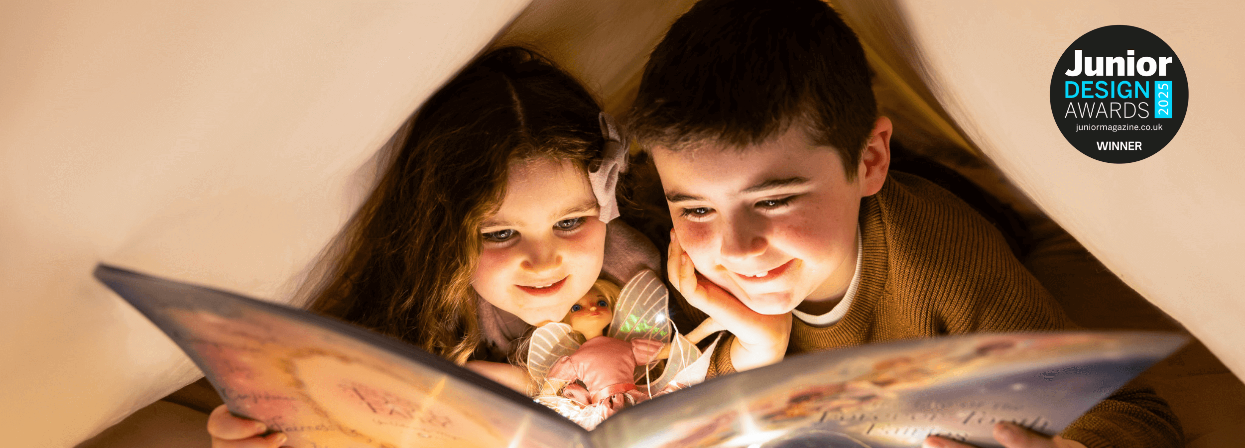 Two children reading a book together under a blanket, with a Junior Design Awards badge in the corner.